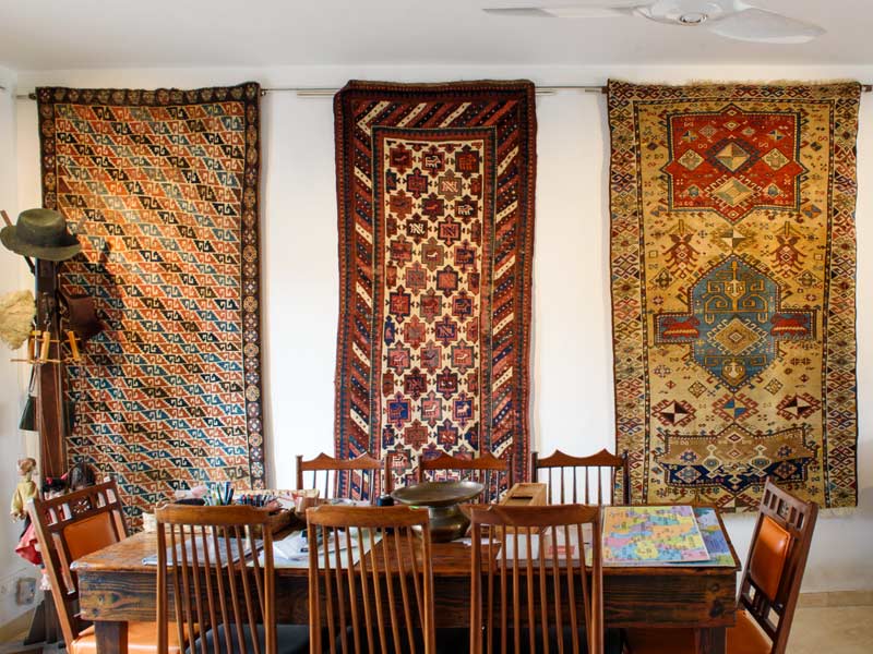 A room features a wooden dining table & chairs, set against a backdrop of colourful, intricately patterned rugs and folk art on the walls - Beautiful Homes